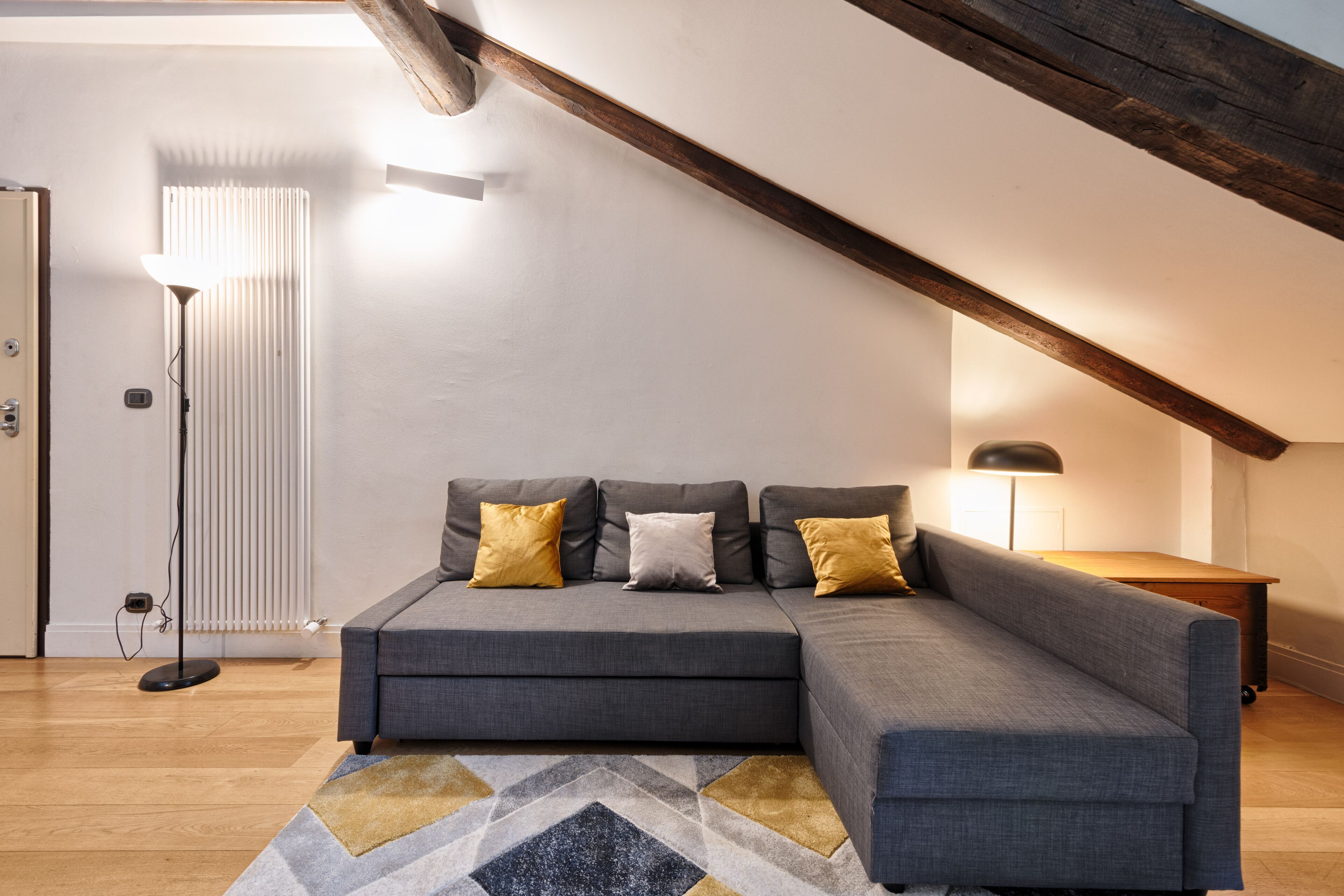 Un salon moderne avec un canapé d’angle gris, un tapis à motifs et une poutre en bois au plafond.