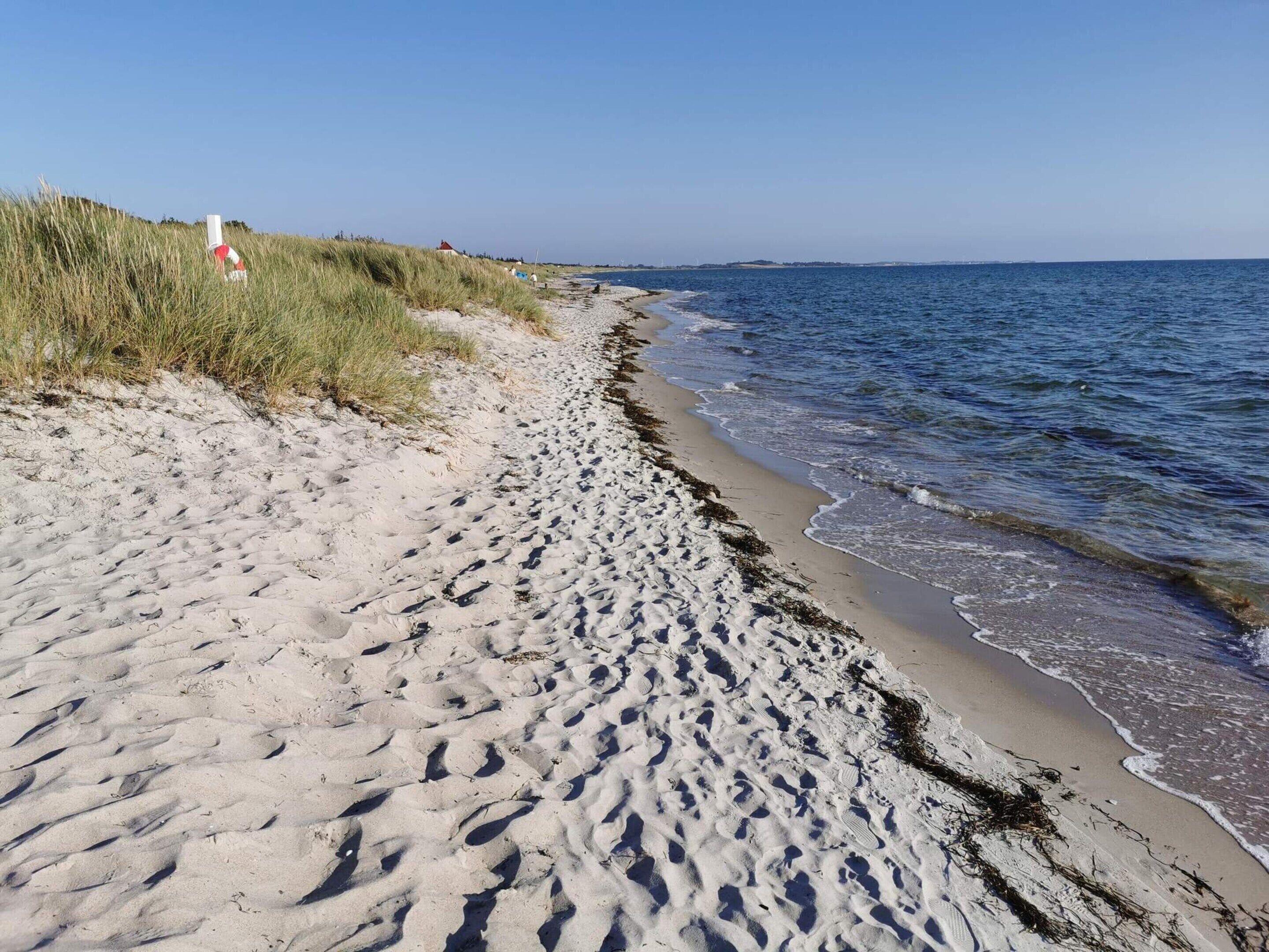 A sandy beach with footprints leading to the water, a grassy dune, and a red and white structure in the distance.