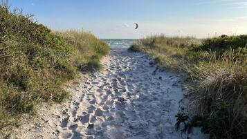 A sandy beach path with footprints leading to the sea, surrounded by grass and vegetation.