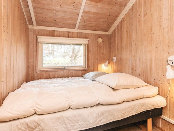 A single bed with a quilted mattress and pillows, a window with a view of trees, and a wall-mounted lamp.