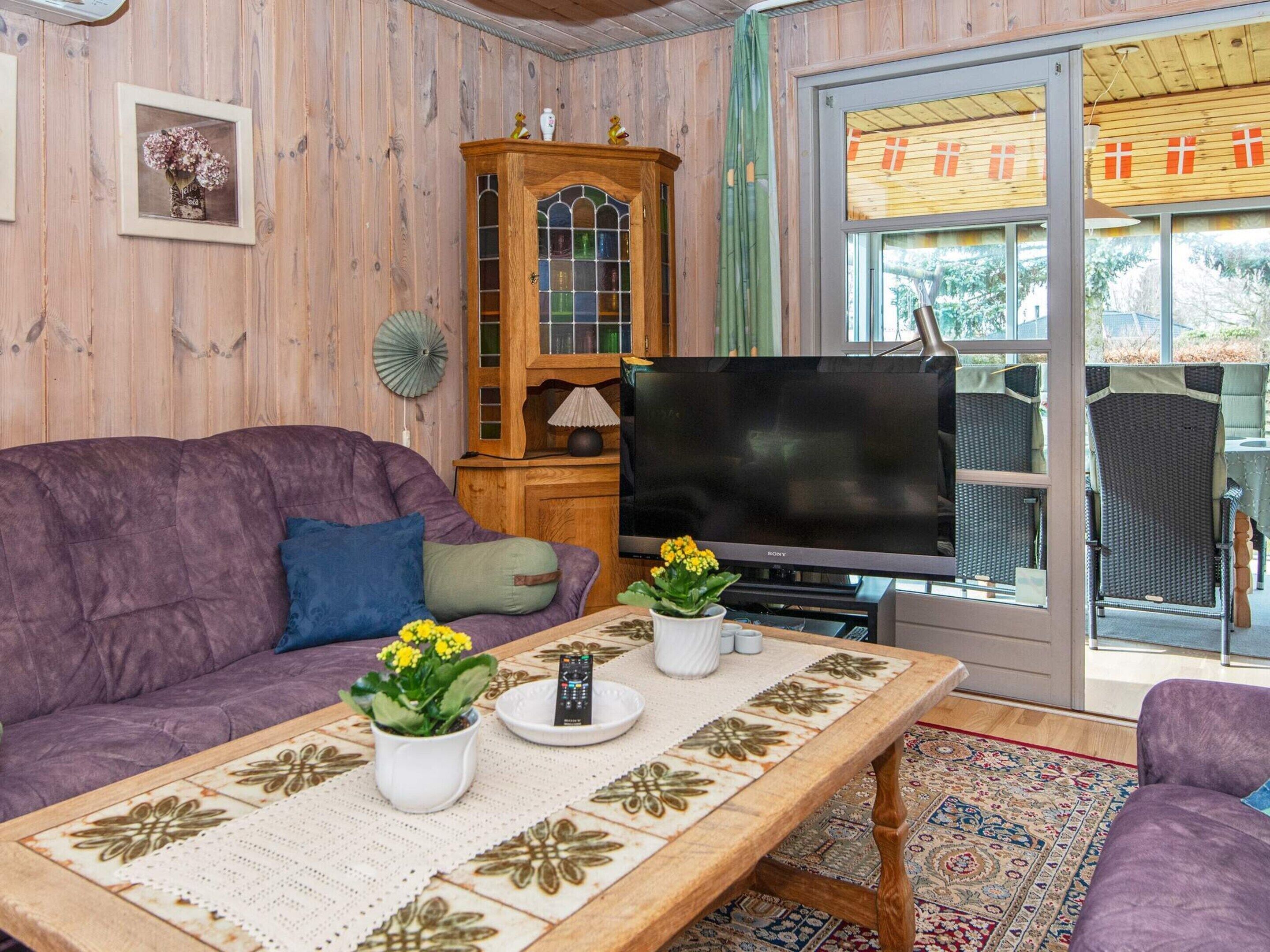 Een woonkamer met een paarse bank, een bijzettafeltje met decoratieve tegels, een houten kast met glazen deuren en een grote flatscreen televisie.