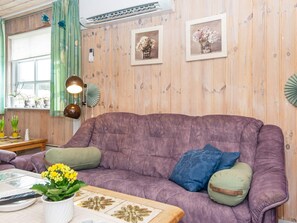 Une pièce avec un canapé violet, une petite table sur laquelle est posé un vase de fleurs jaunes, et une fenêtre ornée de rideaux verts.
