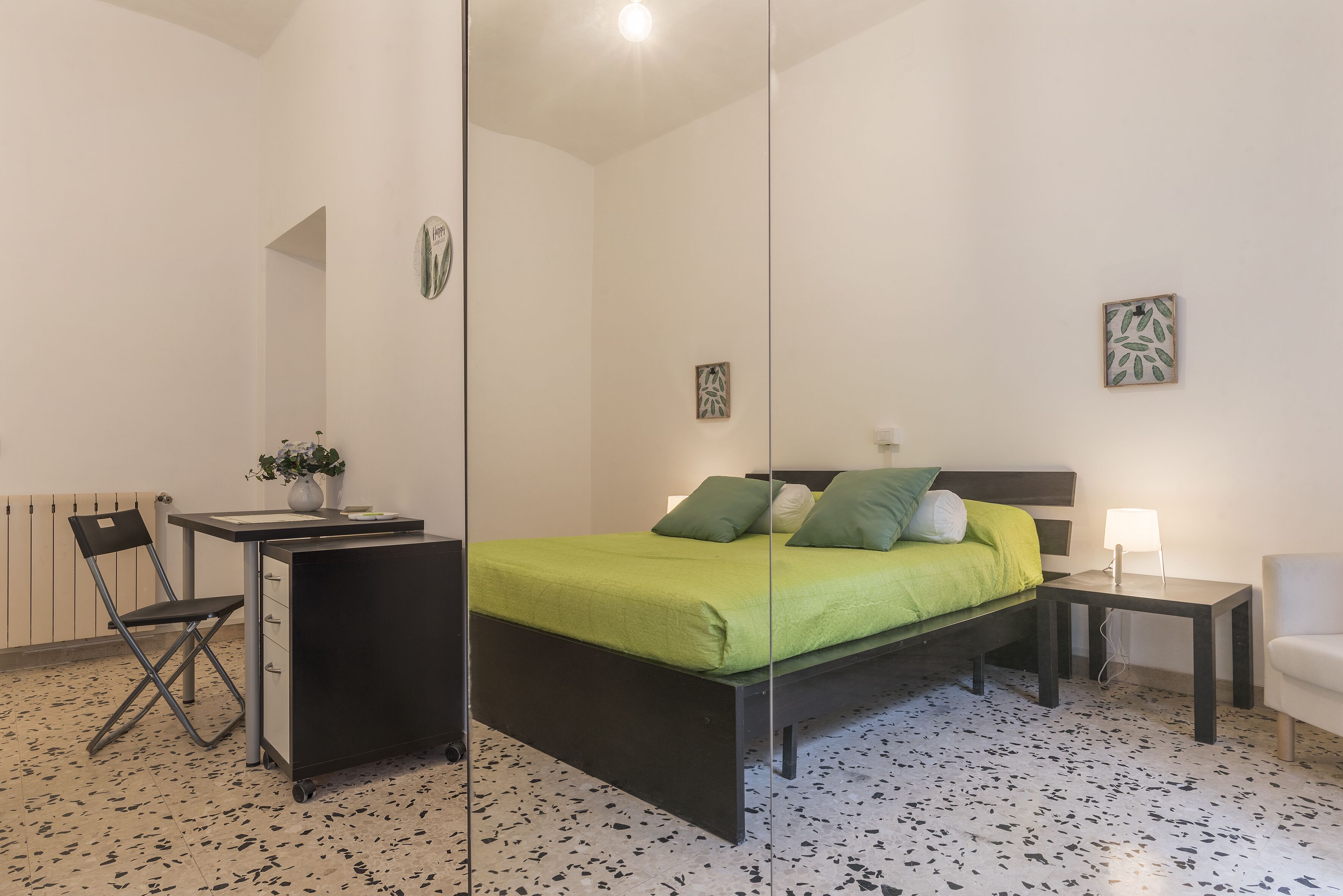 A bedroom with a bed, a desk, a chair, a nightstand, and a wall-mounted mirror.