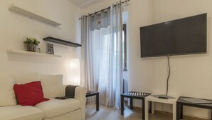 A living room with a white sofa, red pillow, two side tables, a flat-screen TV, and a window with sheer curtains. - Villa Albani Apartment (Rome)