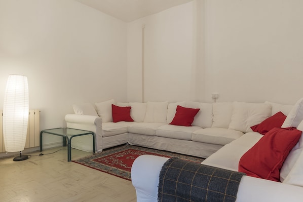 A living room with a white sectional sofa, red cushions, a glass coffee table, and a red and blue patterned rug. - Villa Albani Apartment (Rome)