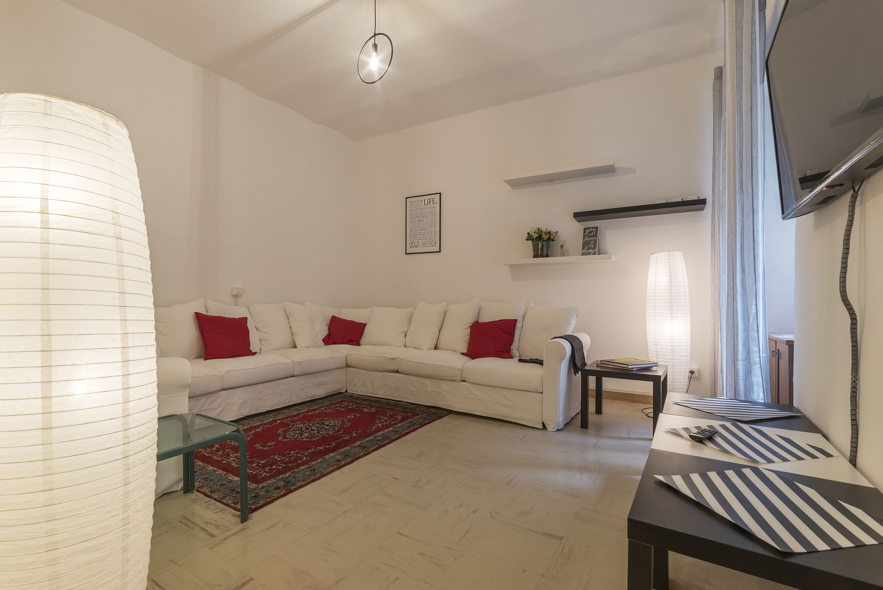 Un salon avec un canapé d’angle blanc, des coussins rouges, une table basse en verre, un tapis à rayures et un téléviseur à écran plat fixé au mur.