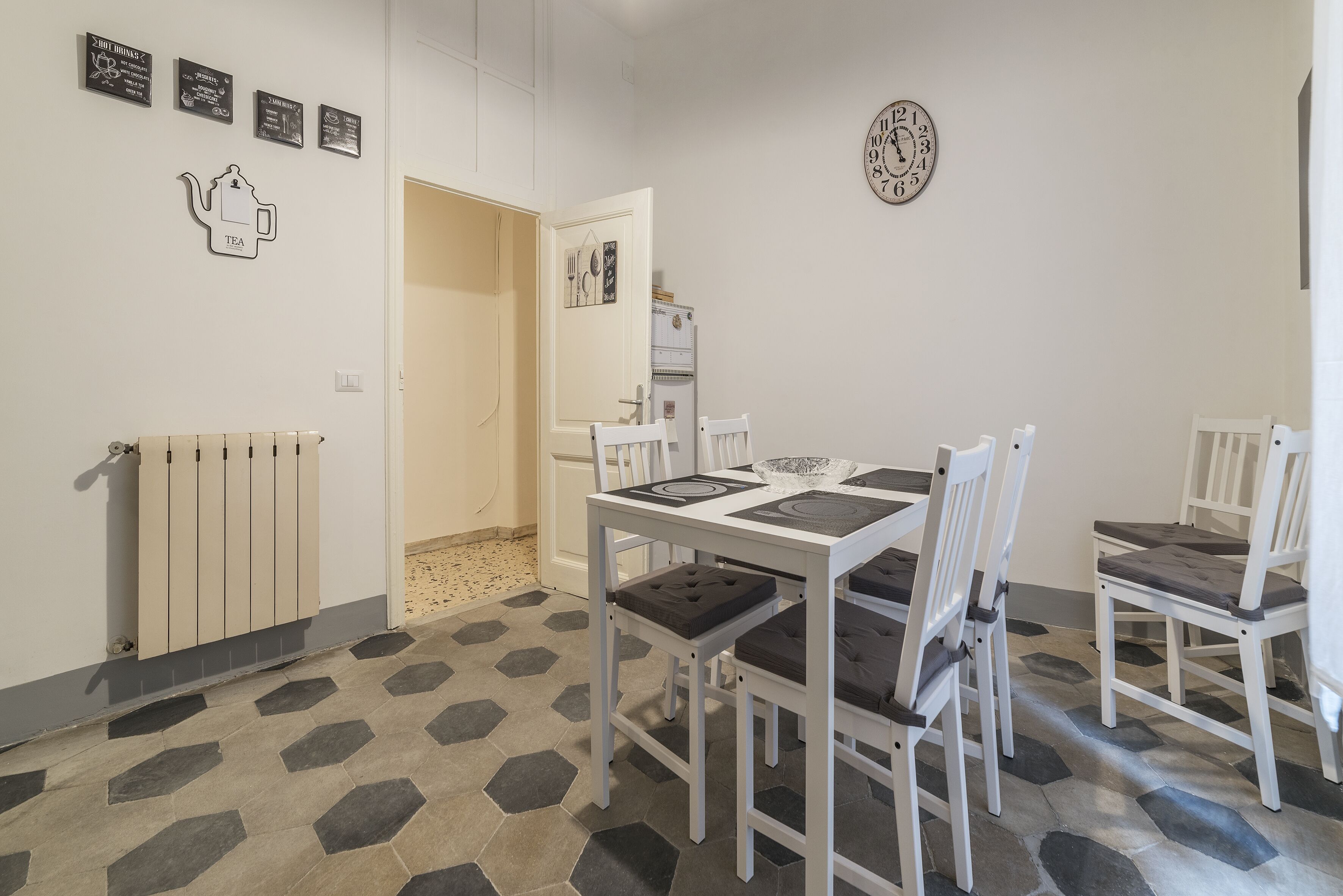 A dining area with a white table and chairs, a hexagonal patterned floor, a radiator, and a wall clock.