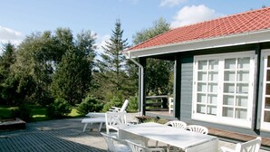 A covered outdoor patio with white plastic furniture, a wooden deck, and a building with a red-tiled roof and large windows. - Spacious Holiday Home With Sauna in Logstor (Logstor)