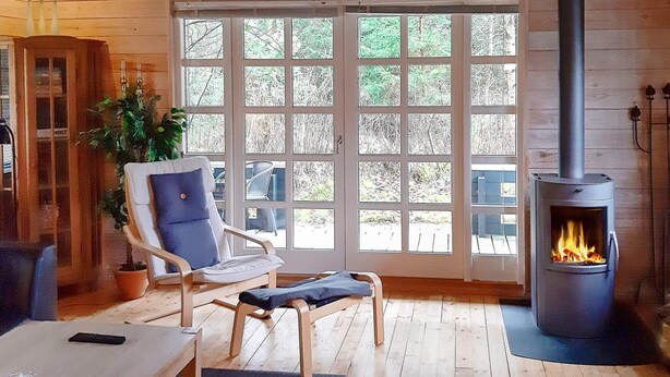 A cozy cabin interior with a large window, a fireplace, wooden furniture, and a potted plant. - Spacious Holiday Home With Sauna in Logstor (Logstor)