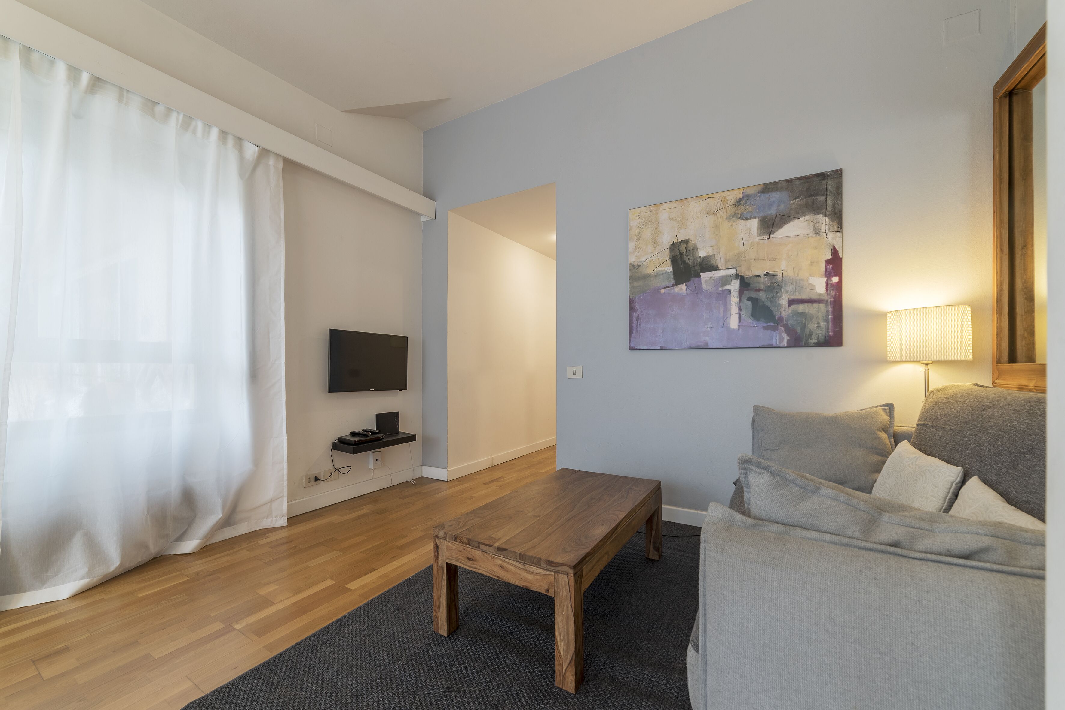 A living room with a wooden coffee table, a grey sofa, a painting on the wall, and a TV on a stand.