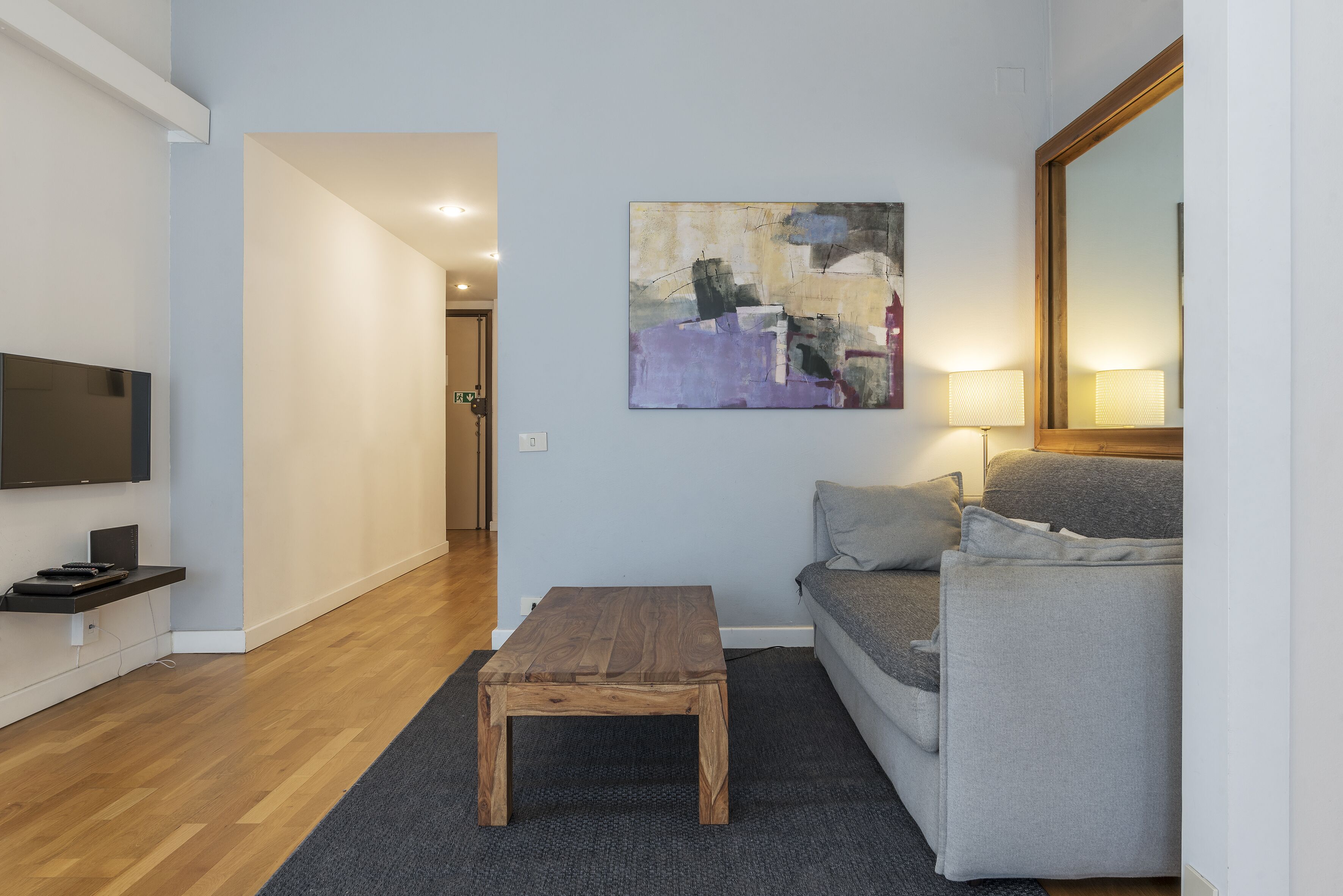 A modern living room with a wooden coffee table, grey sofa, and a painting on the wall.