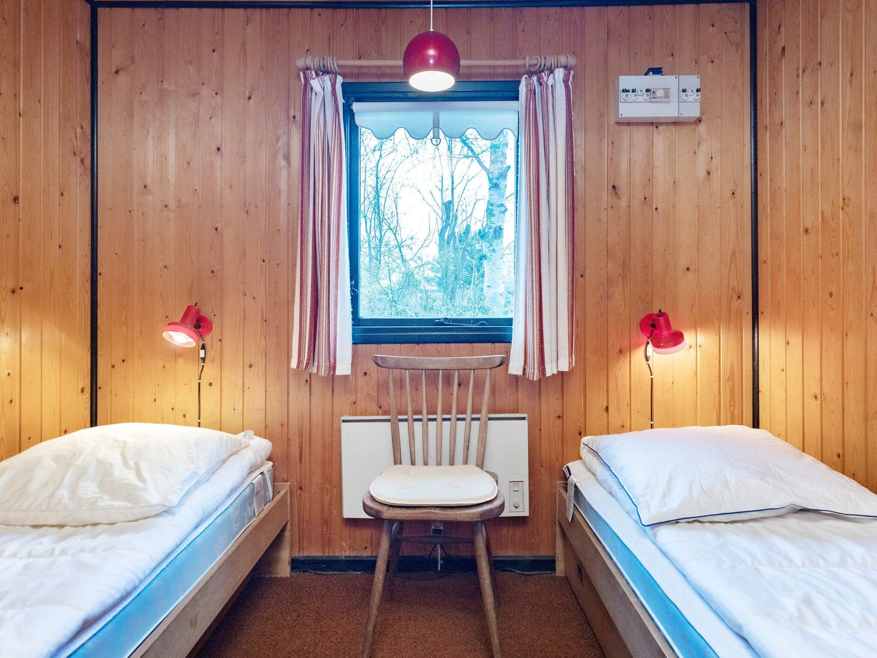 Ein kleines Zimmer mit zwei Betten, einem Sessel, einem Fenster mit Vorhängen und einem fest installierten Heizkörper.