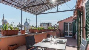 An outdoor patio with a table set for two, surrounded by potted plants, and overlooking a cityscape with historical buildings. - Piazza Navona Panoramic Penthouse (Rome)