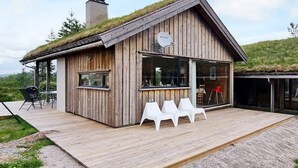 A wooden cabin with a thatched roof, a satellite dish, and a deck with white chairs. - 8 Person Holiday Home in Aseral (Aseral)