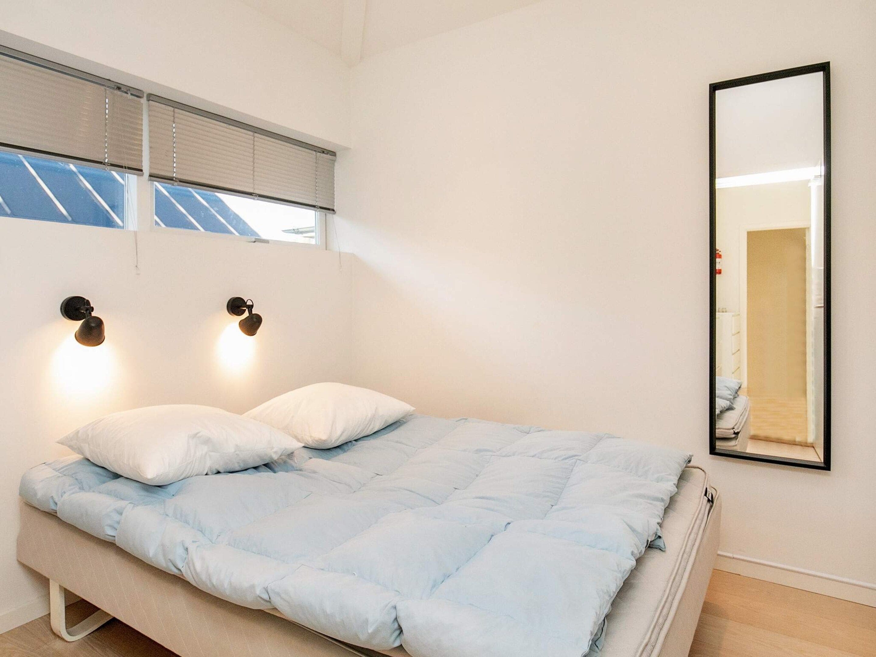 A bedroom with a bed, two wall-mounted lights, a window with blinds, and a mirror on the wall.