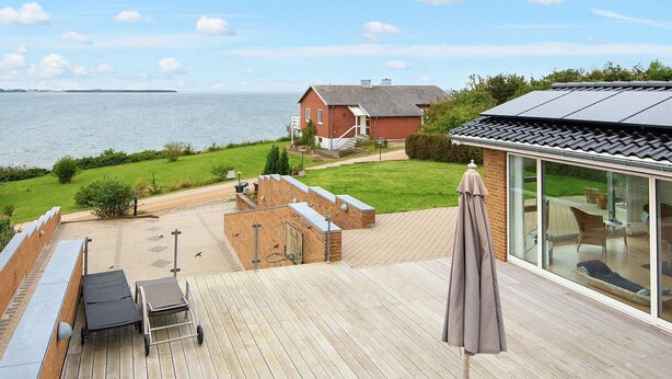 Una terraza de madera con muebles de exterior, una sala con paredes de vidrio que ofrece vistas al mar, y una casa de ladrillo al fondo.