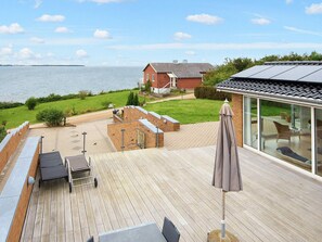 A wooden deck with outdoor furniture, a glass-enclosed room with a view of the sea, and a brick house in the background.