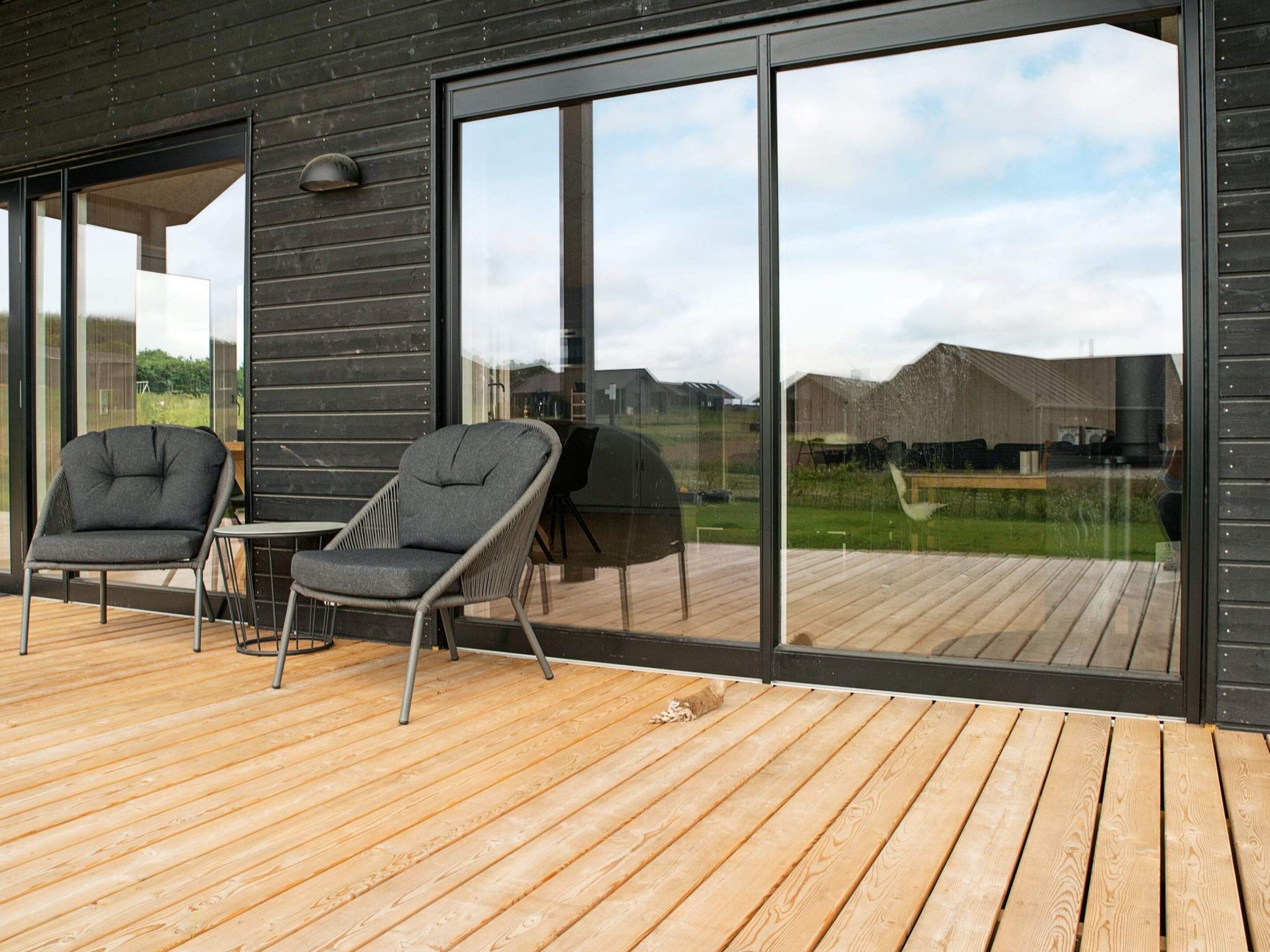 A modern outdoor deck with two chairs and a table, large sliding glass doors, and a view of a residential area.