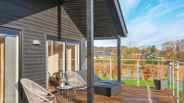 Une terrasse extérieure moderne avec un plancher en bois, deux fauteuils en osier, une petite table et un canapé d’extérieur noir.