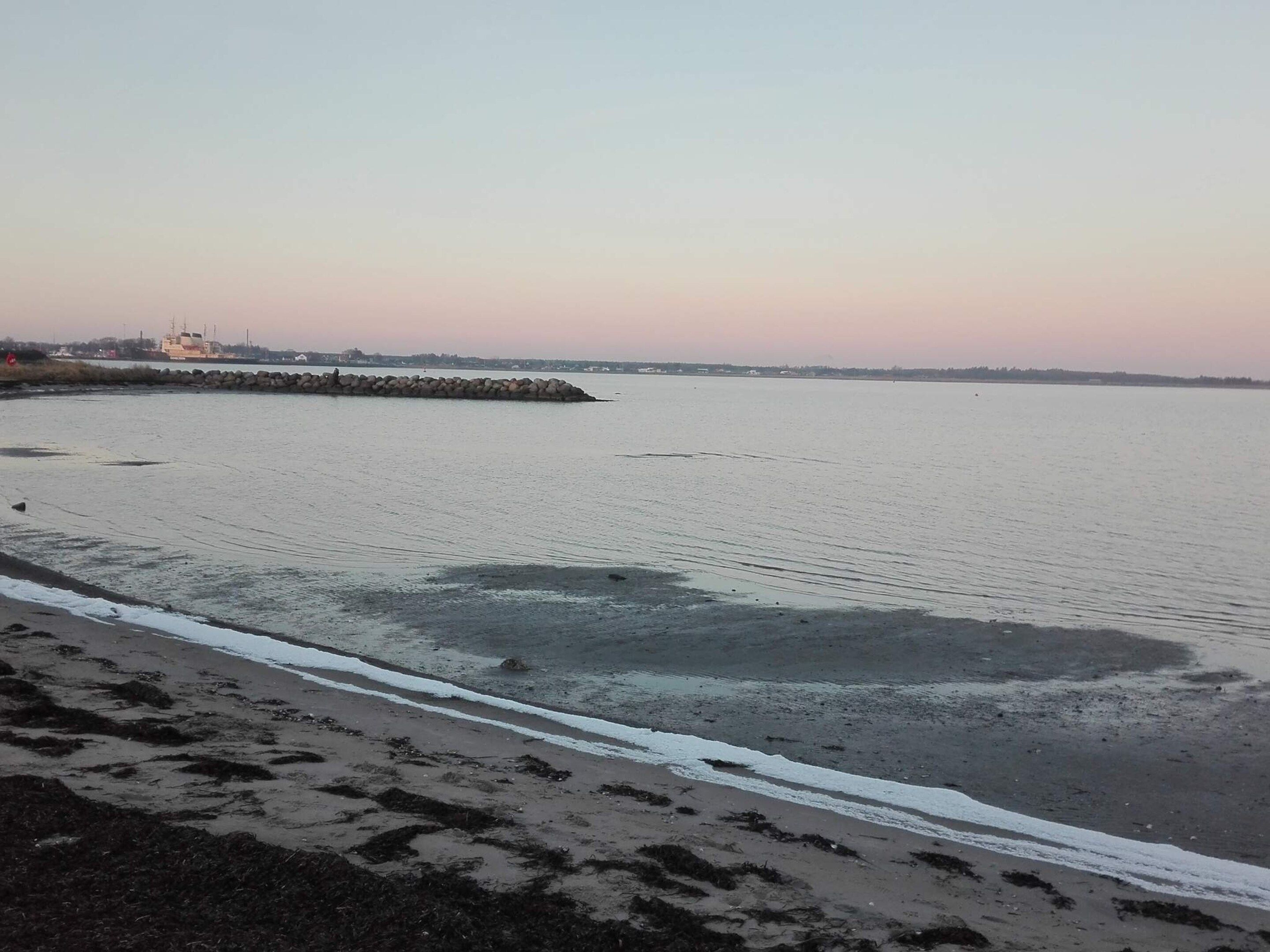 A beach with a stone breakwater, calm water, and a distant shoreline.