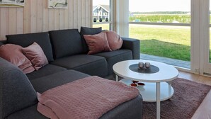 Ein modernes Wohnzimmer mit einem grauen Ecksofa, einem runden weißen Couchtisch und Blick auf ein grünes Feld durch ein großes Fenster.