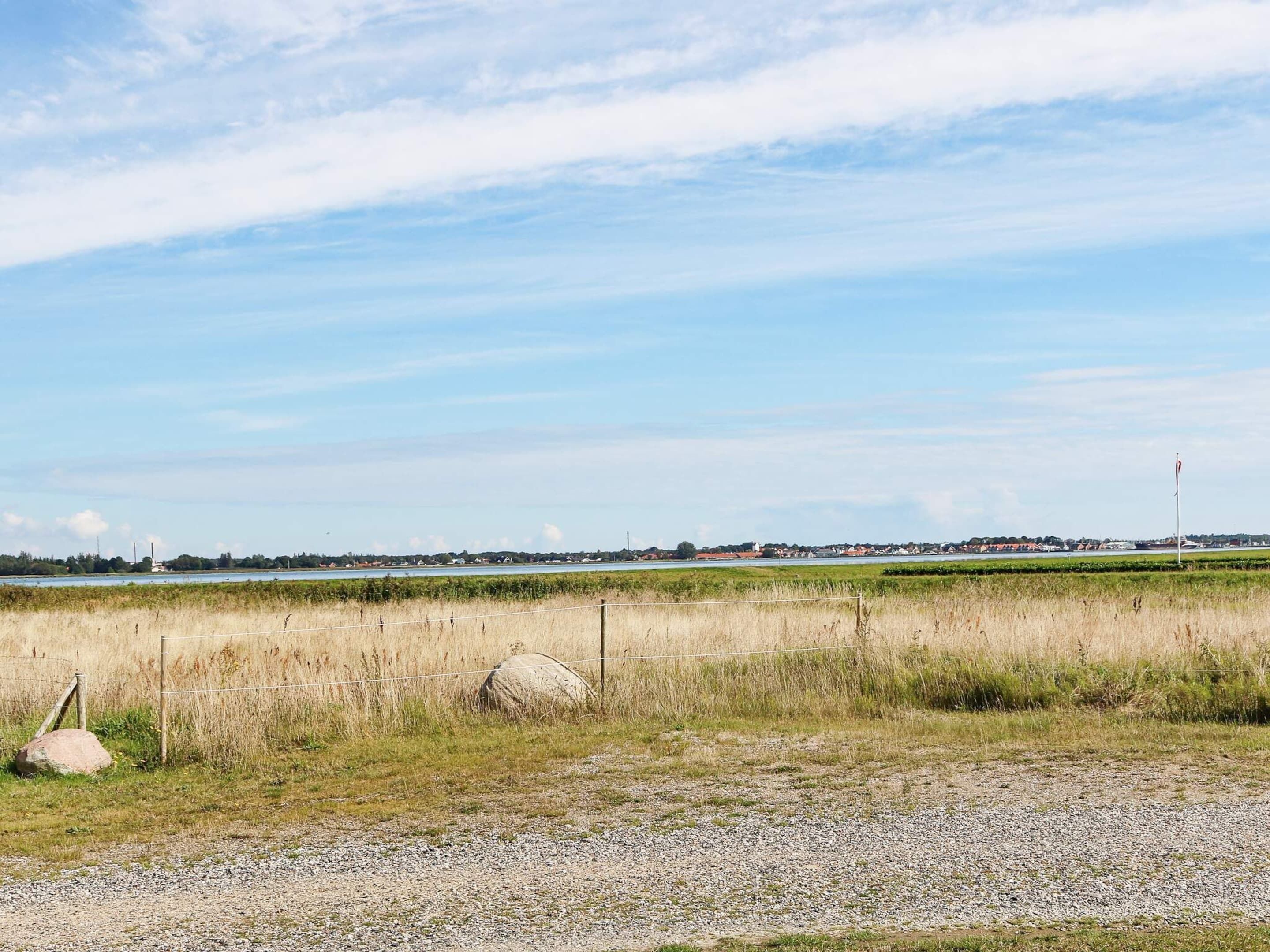 Ländliche Landschaft mit einem Schotterweg, grünen Feldern und einer entfernten Ansicht von Häusern und einem Gewässer.