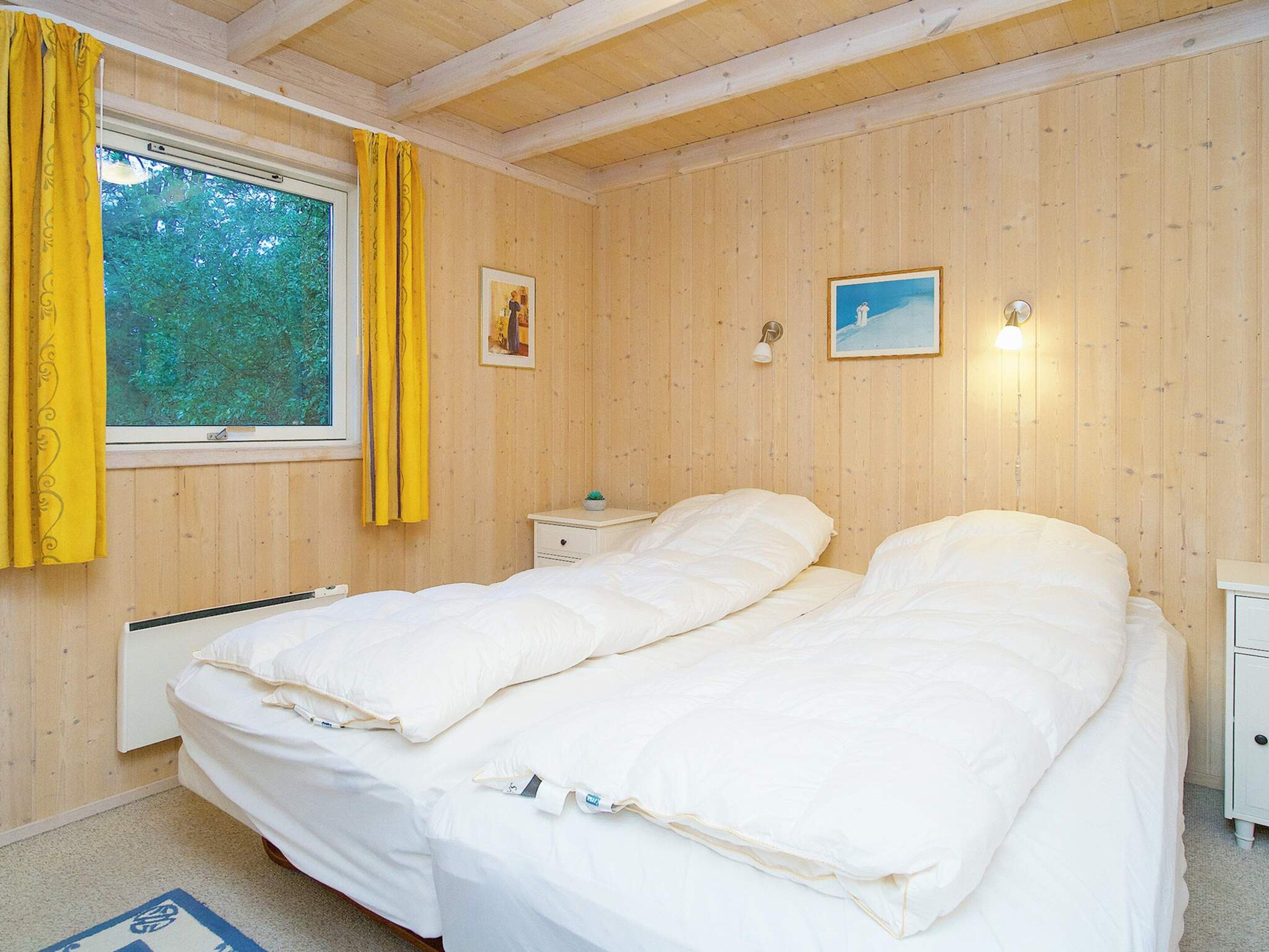 A room with wooden panel walls, a bed with white bedding, a bedside table, a nightstand, a window with curtains, and two wall-mounted lights.