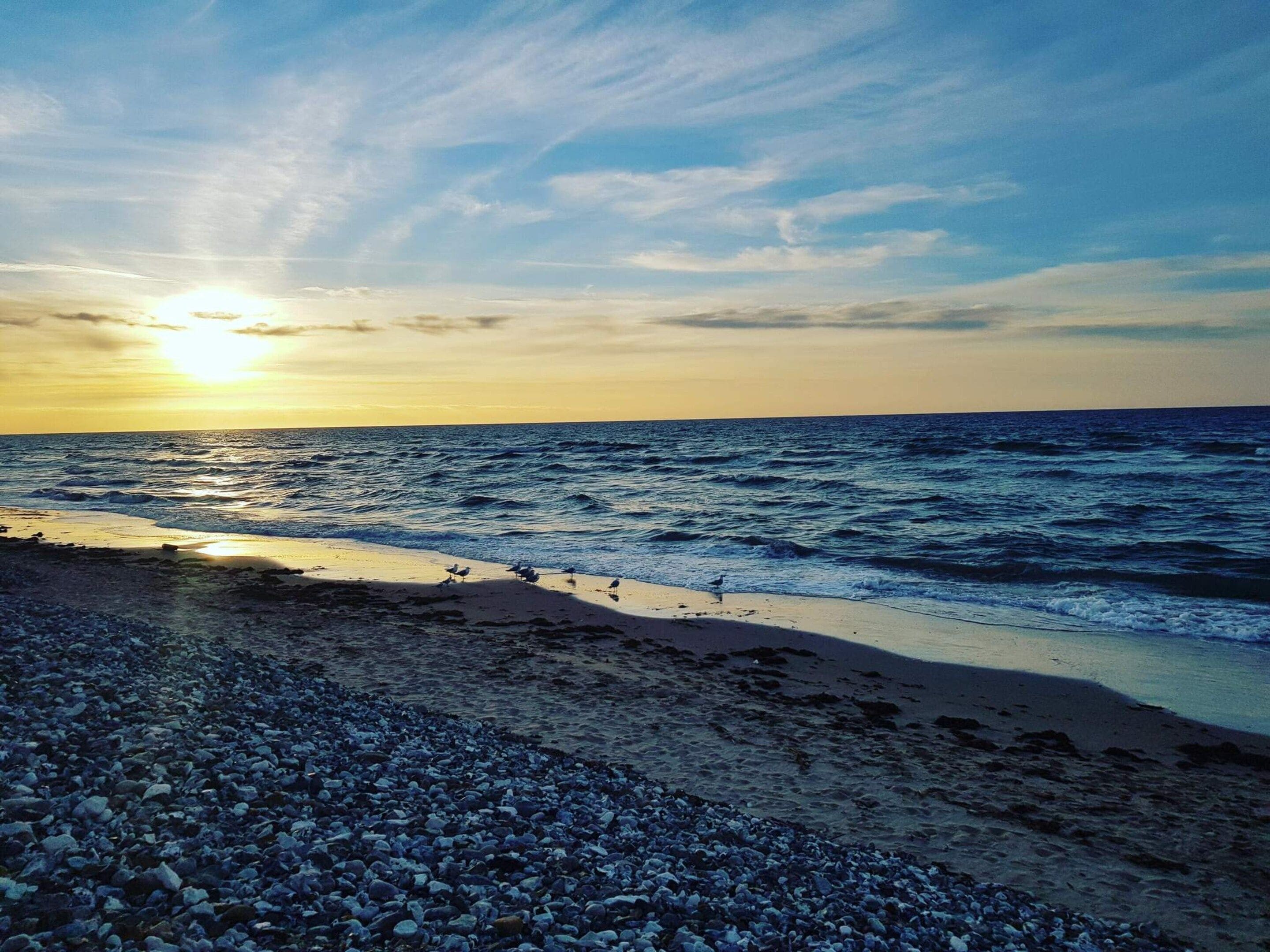 A beach with pebbles and waves, and the sun is setting in the background.