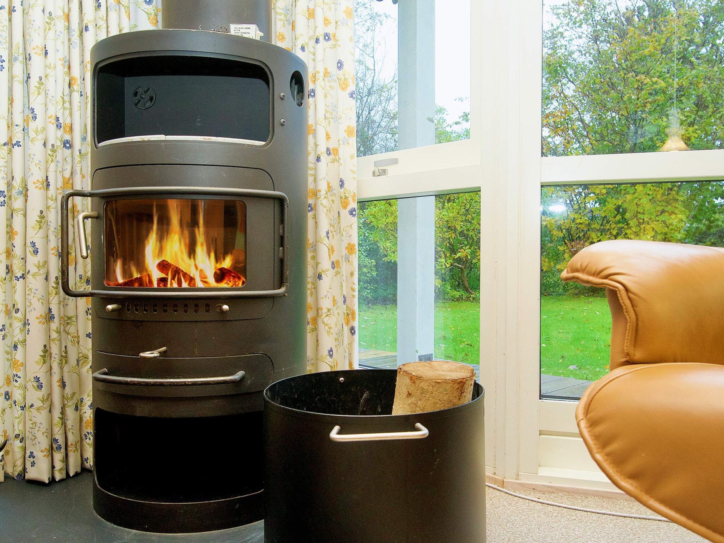 A wood-burning stove with a visible fire, a window with a view of greenery, and a leather chair.
