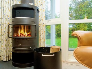 A wood-burning stove with a visible fire, a window with a view of greenery, and a leather chair. - 5 Star Holiday Home in Graested (Graested)