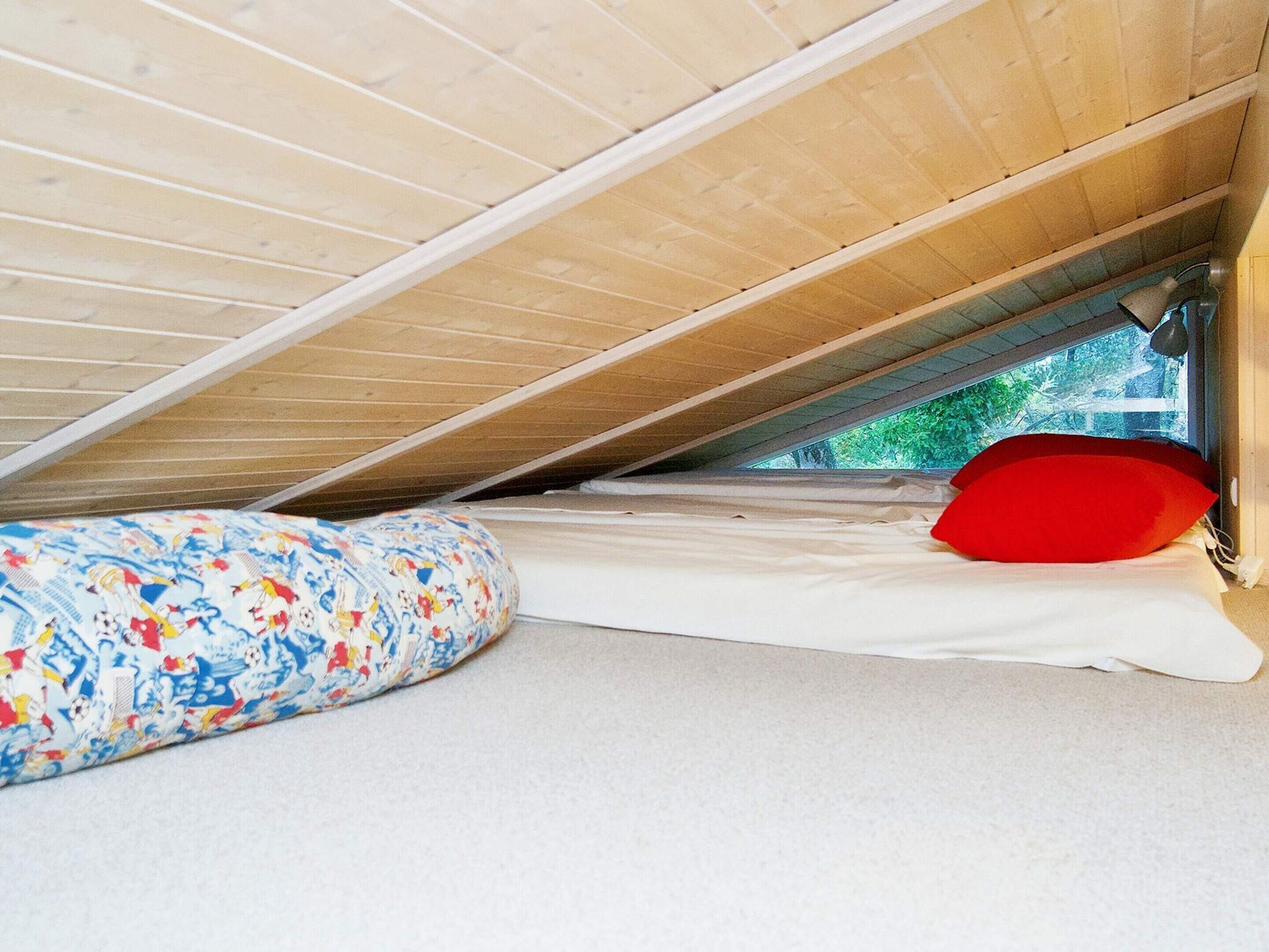 A small, enclosed attic room with a sloped ceiling, a mattress with a colorful blanket, and a red pillow.