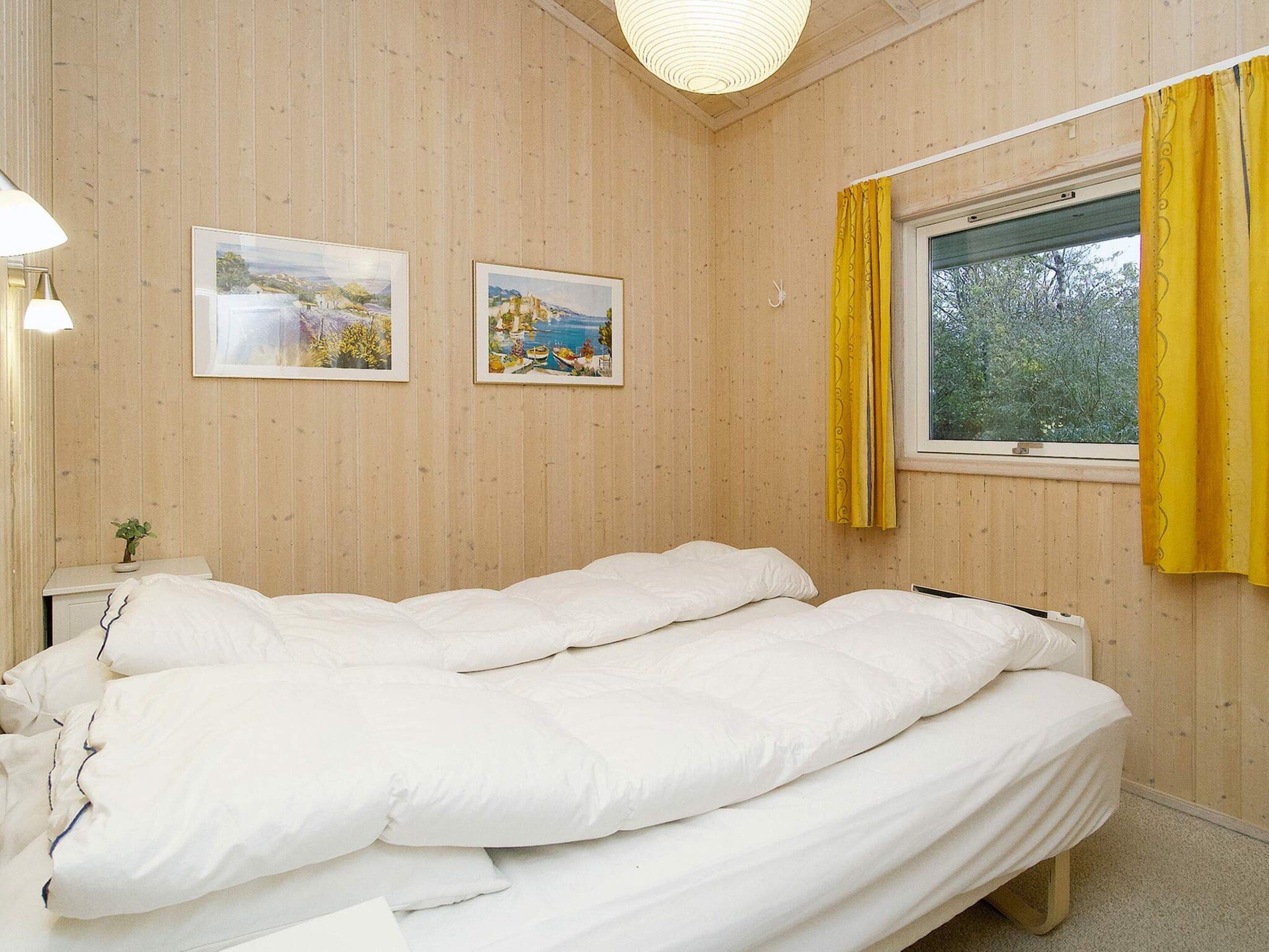 Un dormitorio con cama, dos cuadros en la pared, una mesita de noche con lámpara y una ventana con cortinas.
