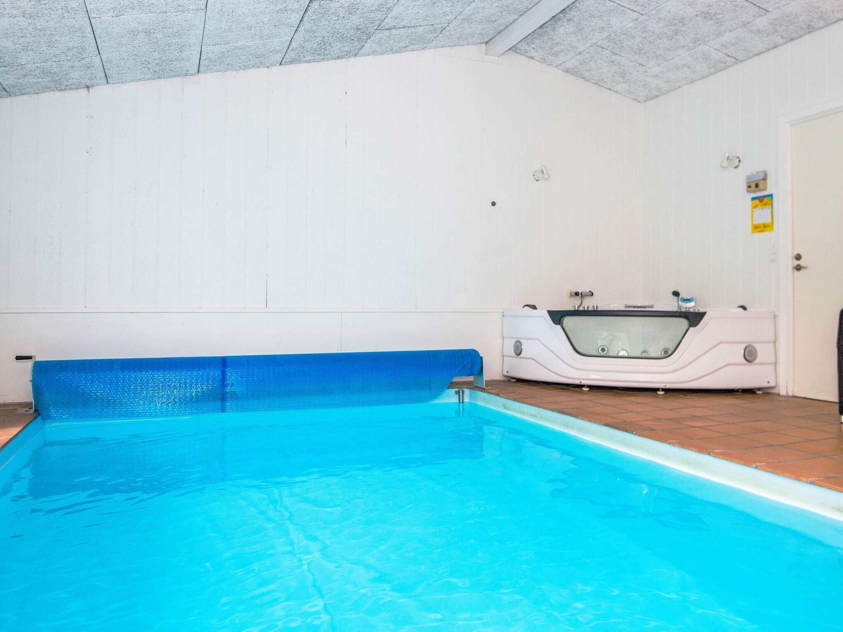 An indoor swimming pool with a blue edge, a jacuzzi, and white tiled walls.