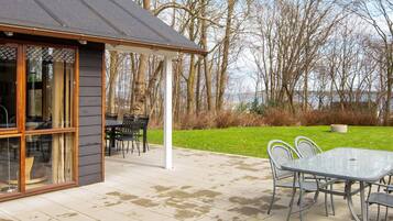 Une terrasse couverte avec une table et des chaises, une entrée avec une porte vitrée et une vue sur les arbres et l’eau.