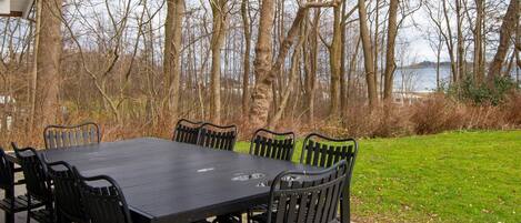 Un patio al aire libre con una mesa rectangular y sillas, rodeado de árboles y vistas a un cuerpo de agua.