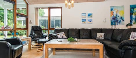 A modern living room with a wooden ceiling, leather sofas, a coffee table, and large windows offering a view of greenery.