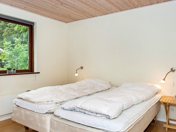 A room with a bed, two bedside lamps, a window with plants, and a wooden ceiling.