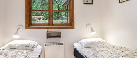 A room with two single beds, a window with a view of greenery, a wall-mounted heater, and two wall-mounted lamps.