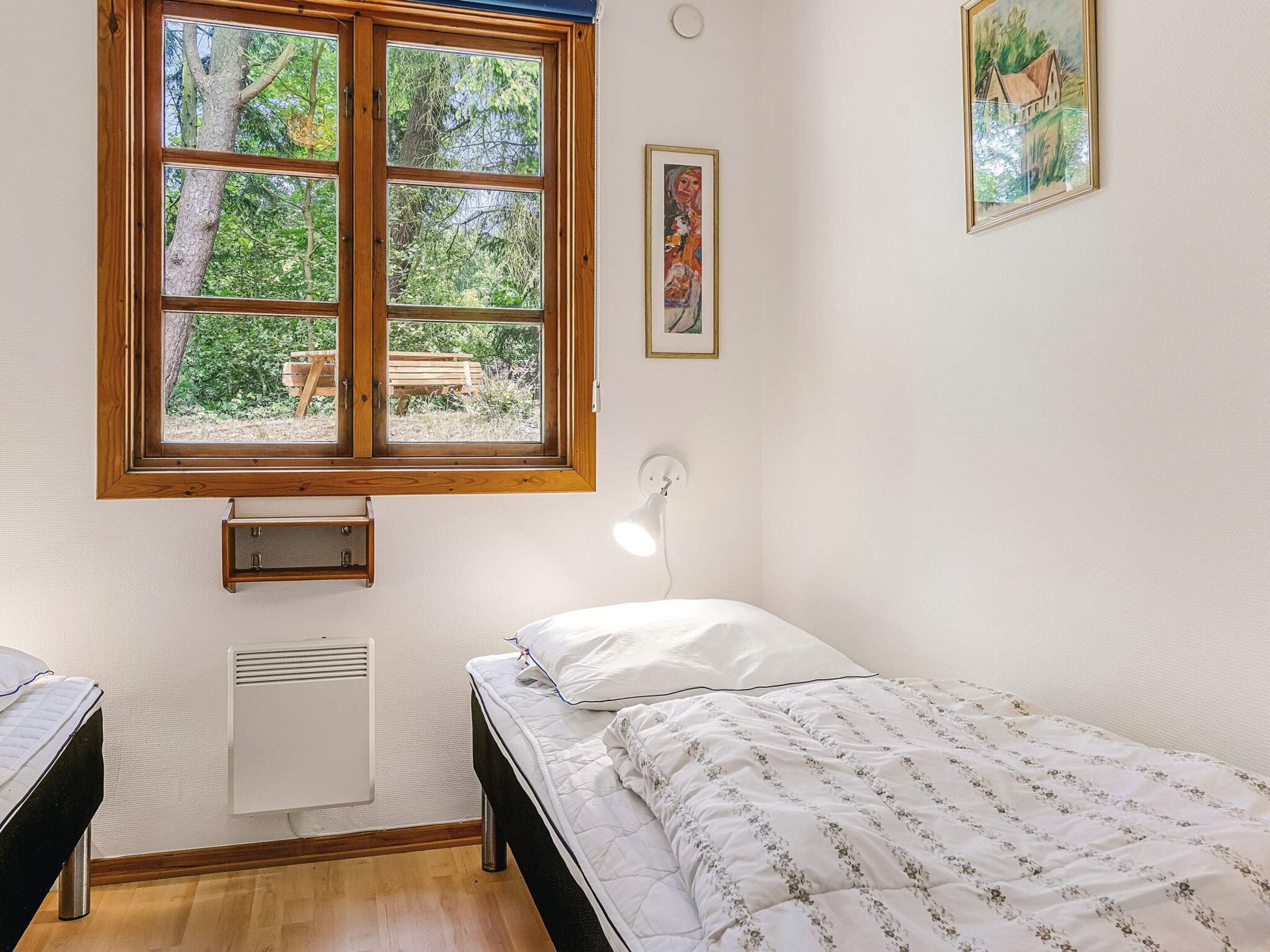 Une chambre avec un lit simple, une fenêtre donnant sur des arbres, un lampadaire fixé au mur et un tableau encadré.