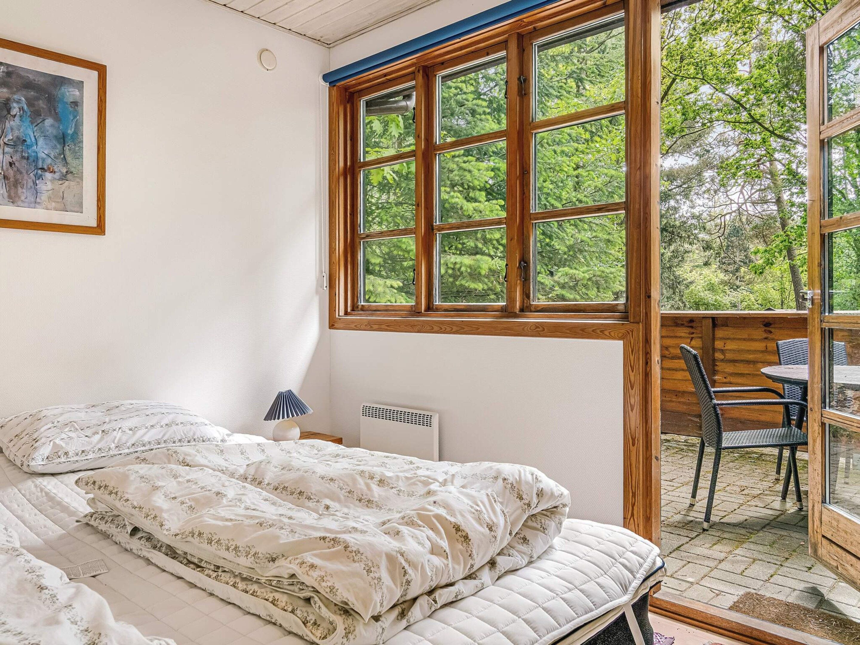 A bedroom with a large window, a bed with floral bedding, a bedside lamp, and a view of the outdoors.