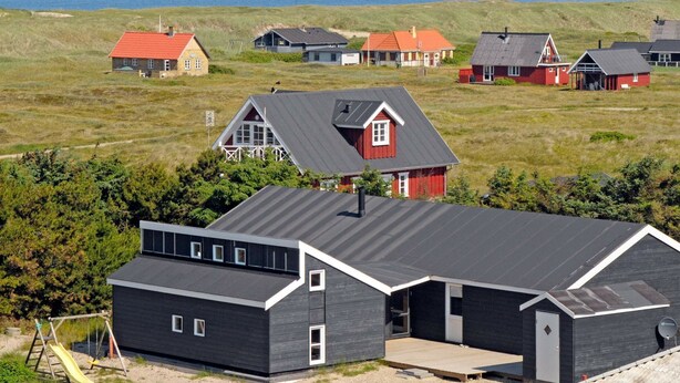 A modern house with a dark exterior and a red roof, situated on a grassy hill overlooking the sea. - Spacious Cottage in Harboøre With Sauna (Harboore)