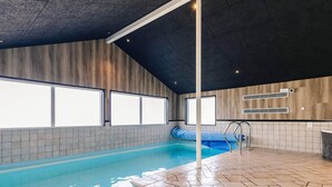 Una piscina interior con paredes de azulejos, techo inclinado y amplia vista del cielo a través de grandes ventanales.