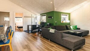 Una sala de estar moderna con un sofá seccional, una mesa de comedor con sillas, un televisor montado en la pared y una pared de acento verde.