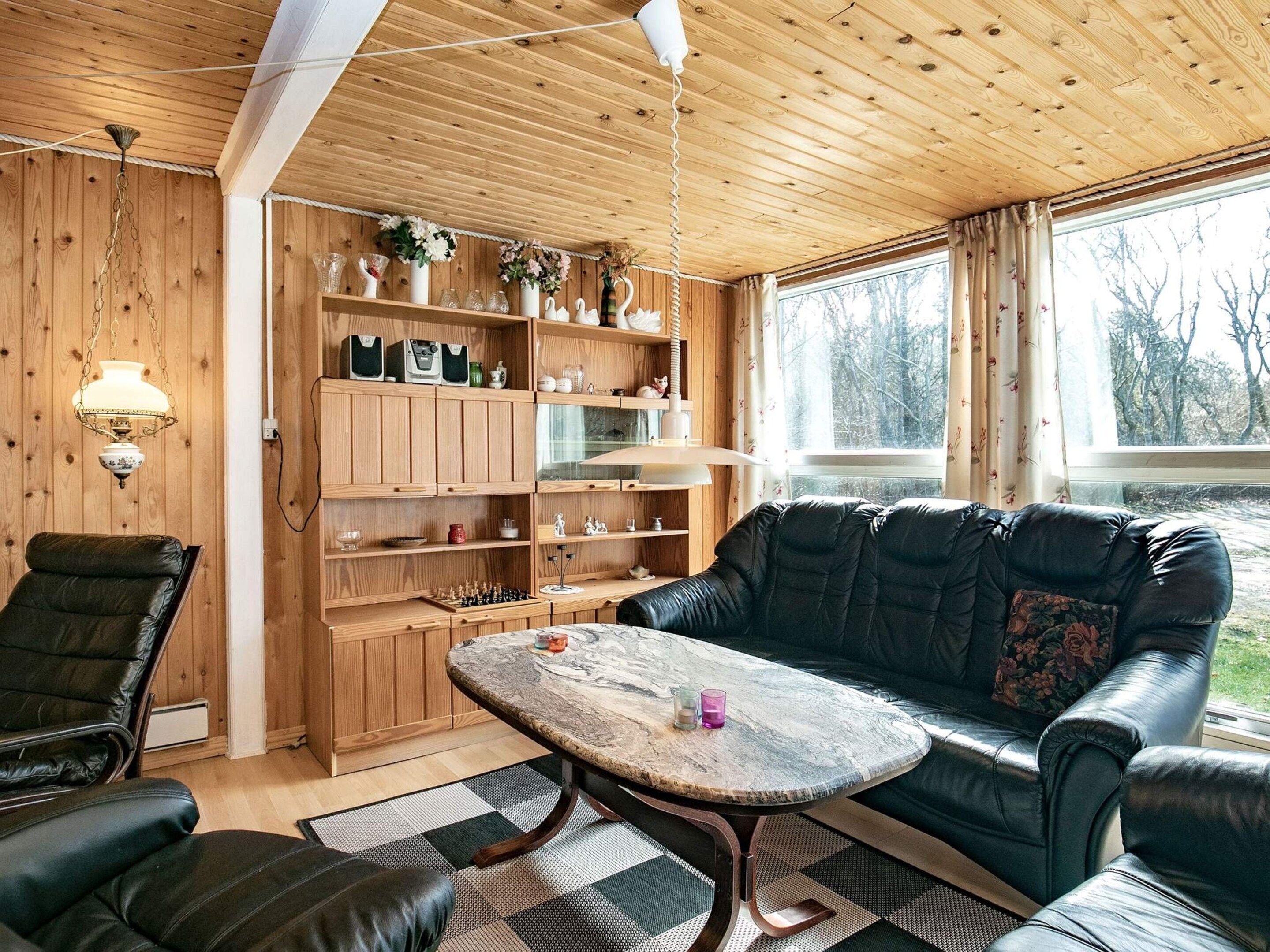 Una habitación acogedora con techo de madera, un sofá de cuero negro, una estantería de madera y un ventanal con vistas a árboles.