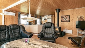 Interior acolhedor de uma cabana com teto de madeira, mesa de pedra com velas, sofás de couro, uma TV e lareira.