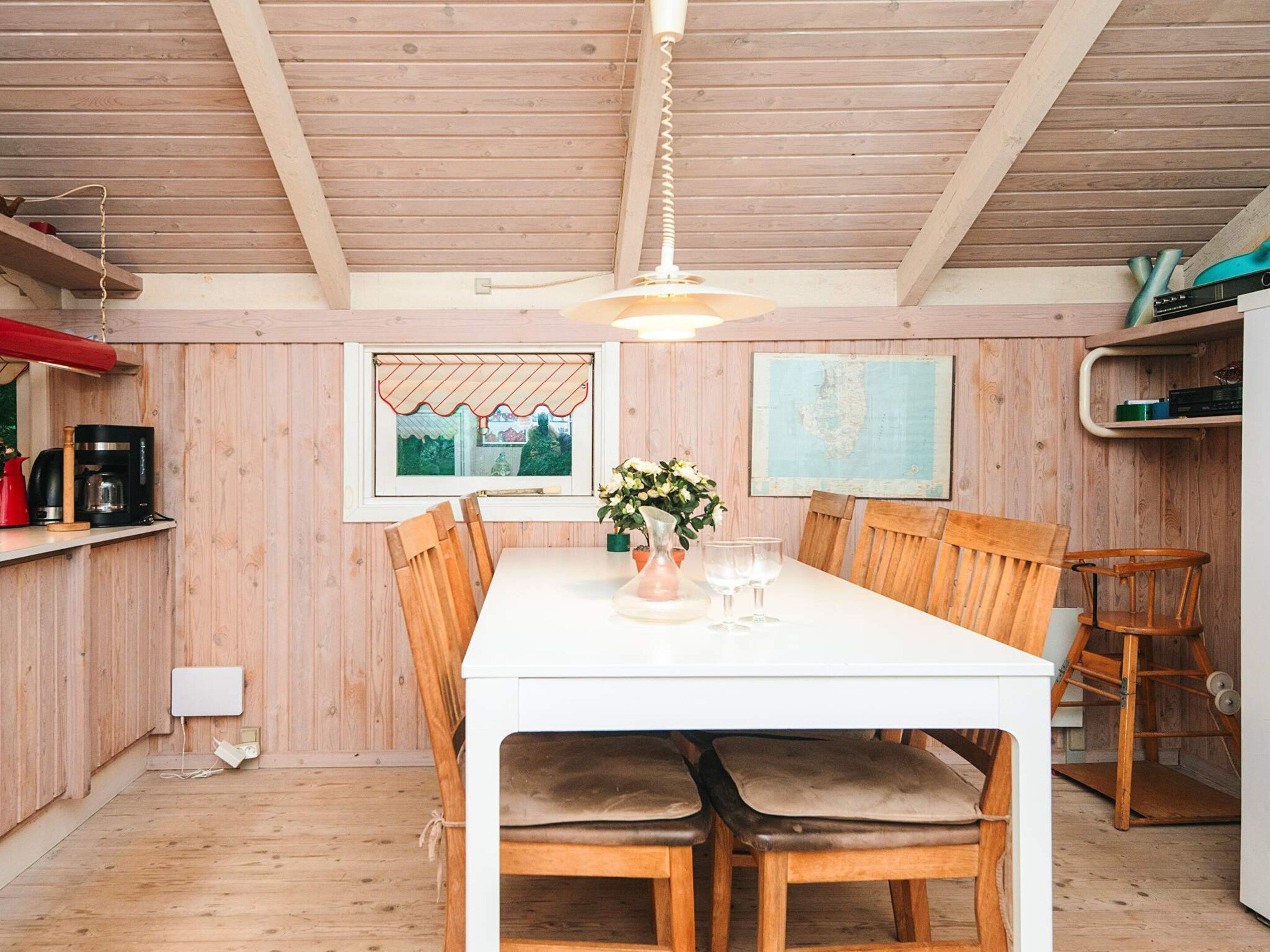 A dining area with a white table, wooden chairs, a hanging light fixture, a window with a view, and a wall-mounted map.