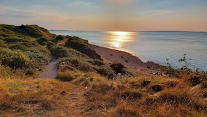 A coastal landscape with a path leading to the beach, a clear sky, and the sun setting over the water.
