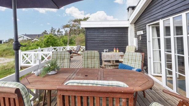 A wooden deck with a table and chairs, a white fence, and a building with a dark exterior and a white window frame.