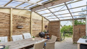 A covered patio area with a wooden table and wicker chairs, a grill, and a view of the outdoors.
