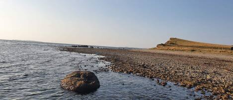 A rocky shoreline with calm water, a large rock in the foreground, and a hill in the background.